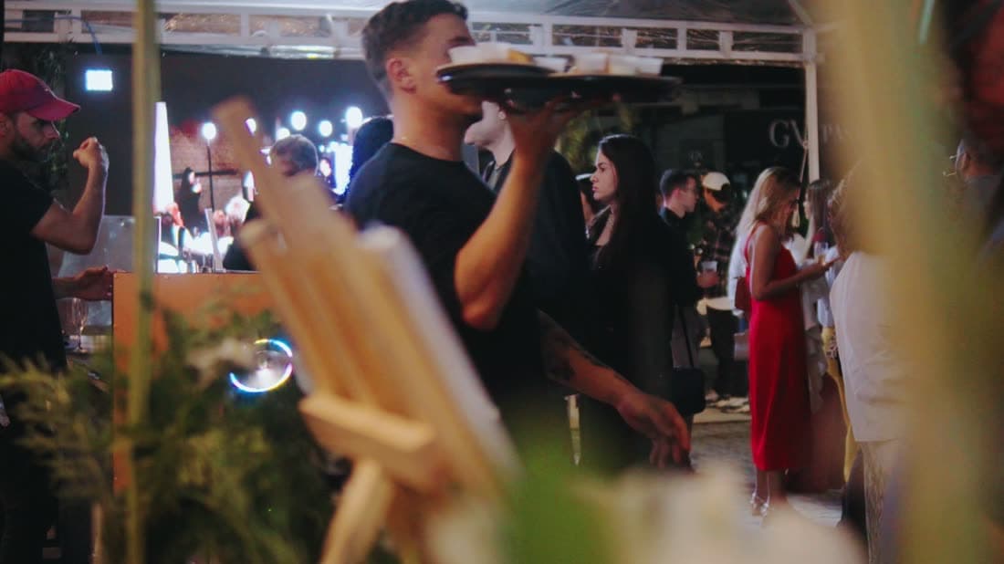 Garçom atravessa o salão com a bandeja erguida durante a inauguração, com a banda ao fundo